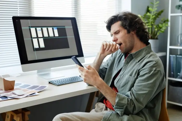 A man yawning while looking at his phone at a cluttered desk with a computer monitor, illustrating the struggle of executive dysfunction in the ADHD vs Laziness debate.