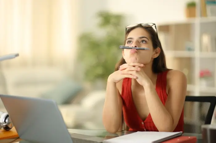A distracted woman balancing a pen on her lip while daydreaming at her desk, illustrating the emotional side of ADHD vs laziness.