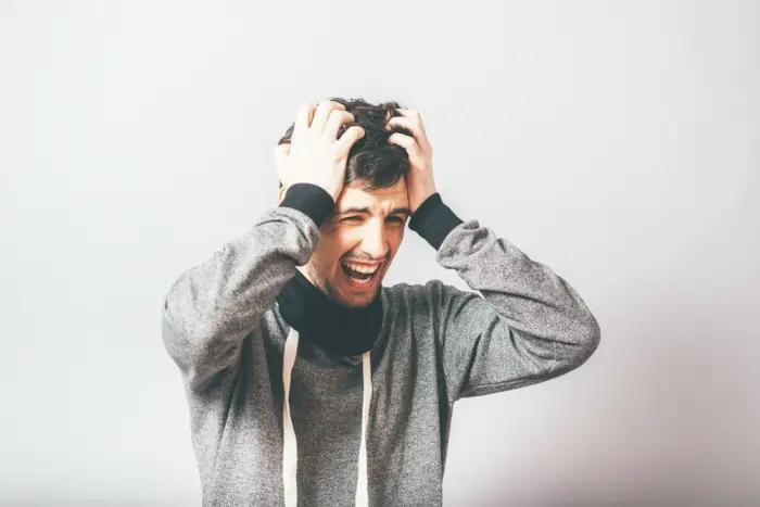 A distressed man in a grey hoodie clutching his hair and shouting with eyes closed, capturing the intense physical restlessness and emotional symptoms of agitated depression.