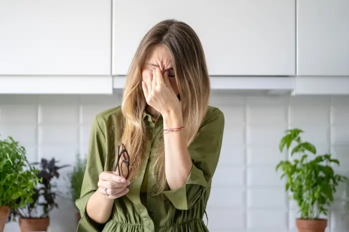 Woman in a kitchen holding glasses and pinching the bridge of her nose, illustrating blurred vision and anxiety.
