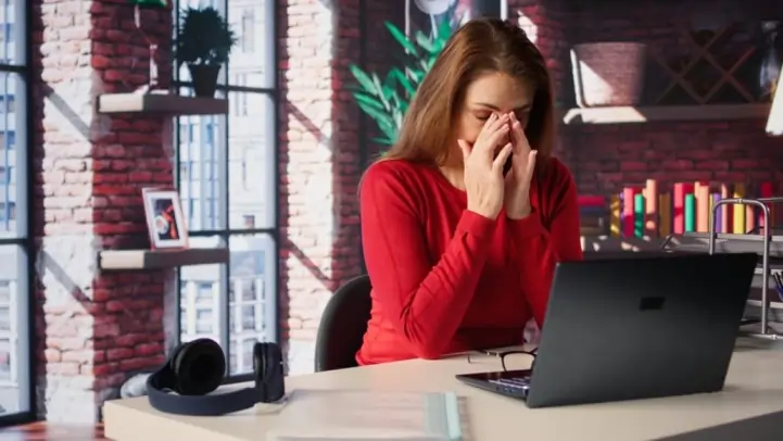 Stressed female holding nose bridge in front of computer showing anxiety causing blurred vision and digital eye strain.