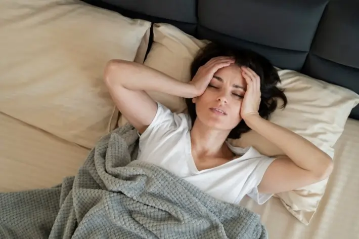 Woman experiencing anxiety during sleep paralysis, holding her head in distress in bed, highlighting the mental and emotional effects of sleep paralysis. Learn about its causes, symptoms, and coping strategies for safer, peaceful sleep.