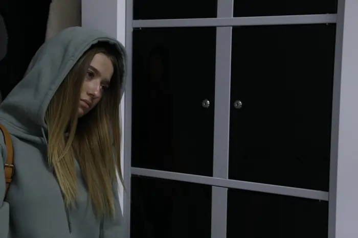 A withdrawn young woman in a hoodie standing in dim light, reflecting the emotional emptiness of apathy and depression.