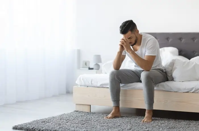 A man sitting on the edge of a bed with his head in his hands, portraying the heavy physical and emotional weight of apathy and depression.