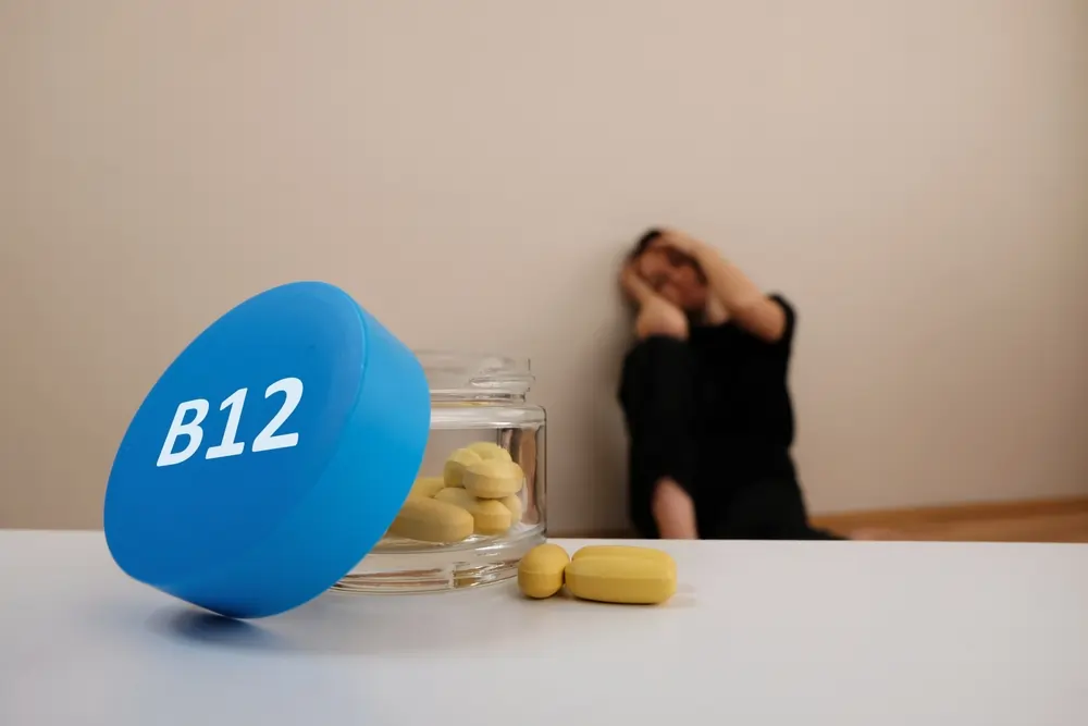 B12 bottle and pills in focus with a blurred person in the background holding their head, depicting anxiety.