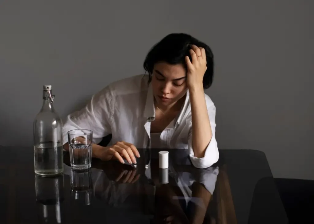 Woman feeling fatigued and anxious sitting at table with pills and water symbolizing B12 deficiency anxiety and mental health imbalance.
