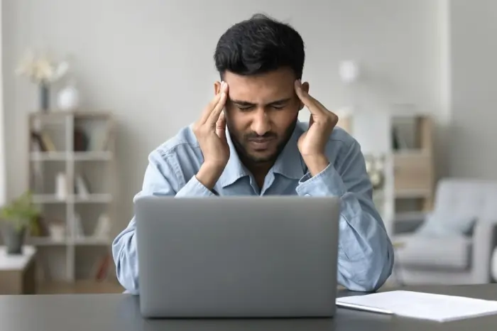 A man sitting at a laptop rubbing his temples in frustration, illustrating the link between depression and memory loss, specifically the difficulty with concentration and cognitive focus during work.