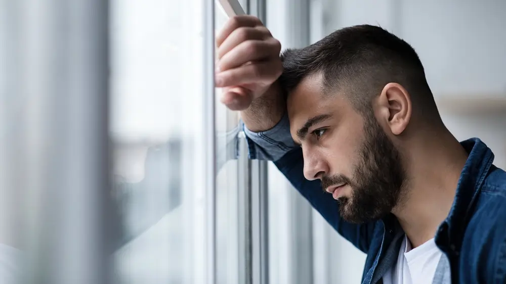 A man leans his head against a window with a somber expression, looking out to visualize isolation depression.