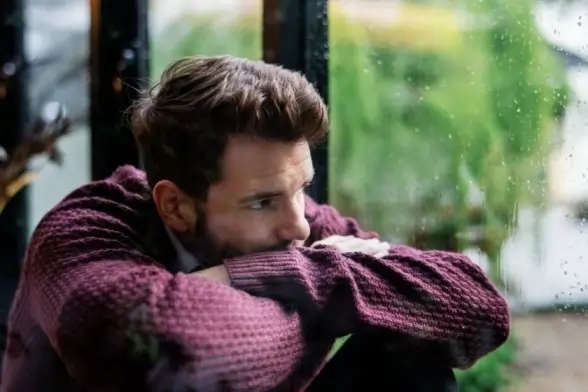 A lone man in a purple sweater gazes through a rain-streaked window, reflecting the silent weight of loneliness and depression.