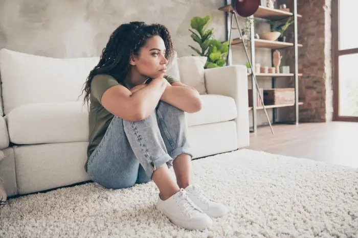 A lonely woman sits on the floor leaning against a white sofa, looking away sadly to represent loneliness depression.