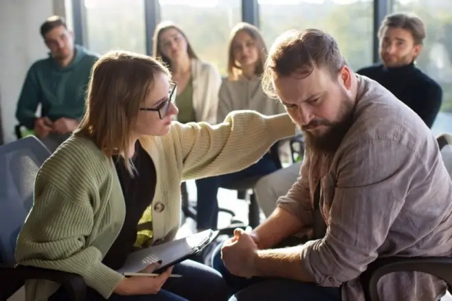 A supportive woman in glasses places a comforting hand on the shoulder of a distressed man looking down in a therapy group setting, illustrating the emotional complexity of why people project feelings onto others.