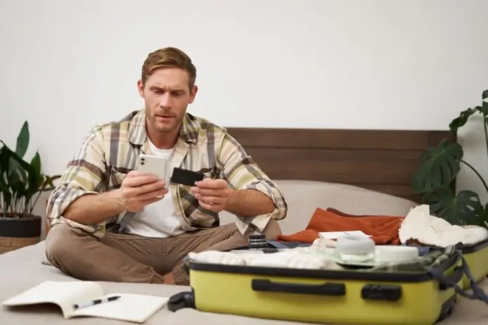 Man sitting on a bed packing a suitcase while checking his phone, looking anxious and stressed before traveling, representing travel anxiety and fear of traveling.
