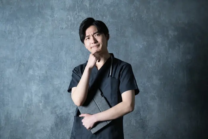 A young professional man in dark scrubs holding a tablet and touching his chin thoughtfully, illustrating deep-seated anxiety about the future.