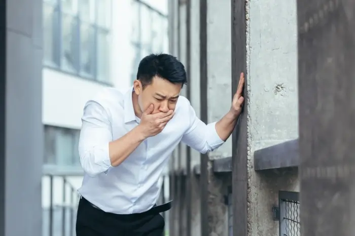 A man in a white shirt leaning against a wall and covering his mouth as if nauseated, illustrating the connection between anxiety and physical ailments like digestive distress.