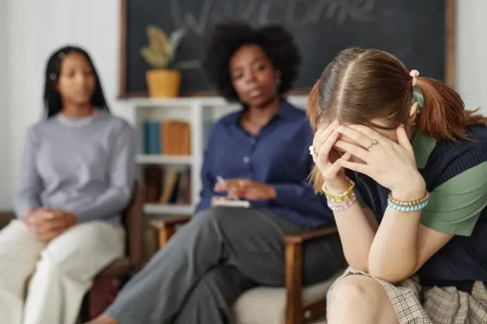 Inhibition and anxiety shown by a young girl covering her face with her hands during a group therapy session or classroom discussion.