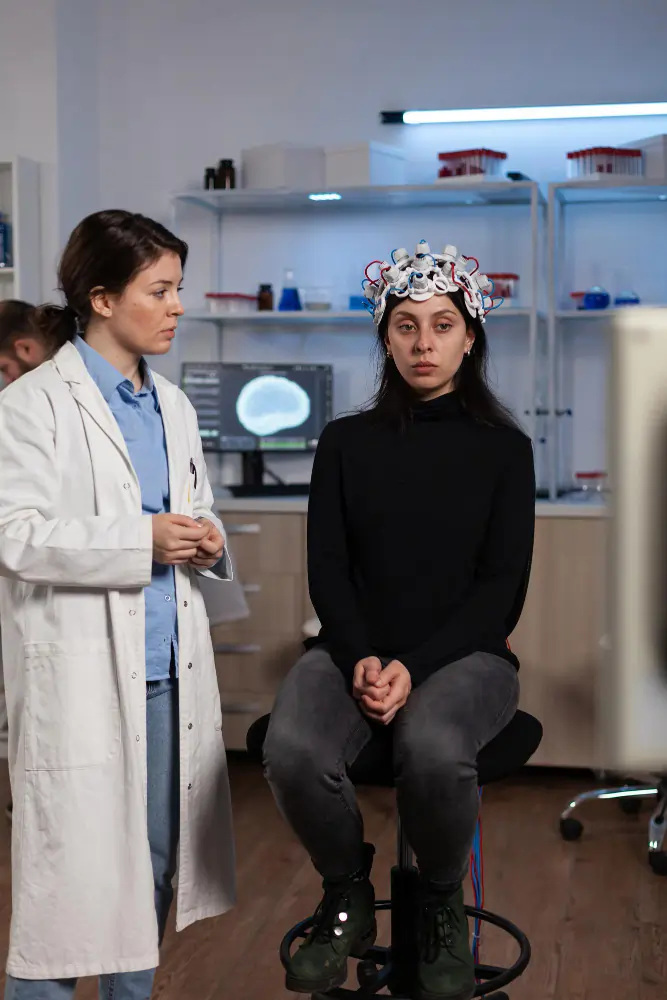 Doctor in a lab coat standing beside a seated woman wearing an EEG cap with electrodes in a medical lab, with a brain scan displayed on a monitor in the background.