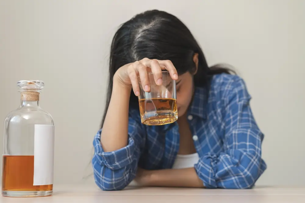 A woman sitting with her head down holding a glass of alcohol, showing emotional distress and struggles with alcohol addiction.