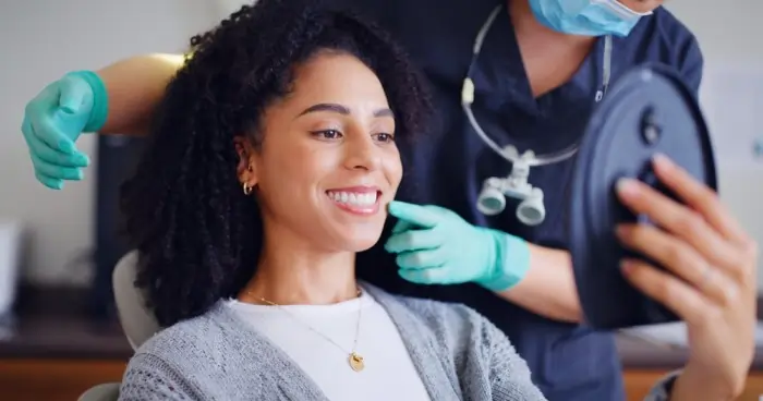 A smiling woman looking in a mirror at her white teeth after a professional dental cleaning procedure at a dentist's office.